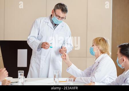 Ricercatore senior in maschera medica mostrando fiala con nuovo vaccino ai suoi colleghi alla conferenza medica Foto Stock