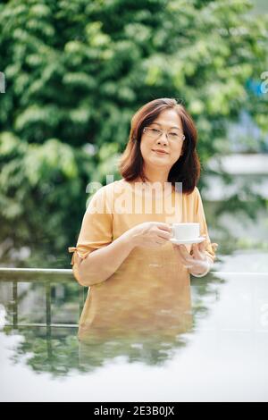 Ritratto di una donna asiatica di età positiva con tazza di caffè in piedi sul balcone e guardando la fotocamera Foto Stock