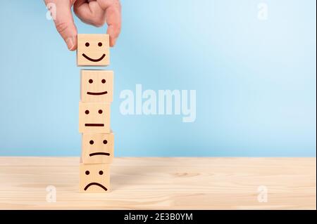 Valutazione del servizio clienti e analisi della soddisfazione concetti. La mano del cliente ha scelto felice faccia sorriso simbolo su blocchi di legno, sfondo blu. cop Foto Stock