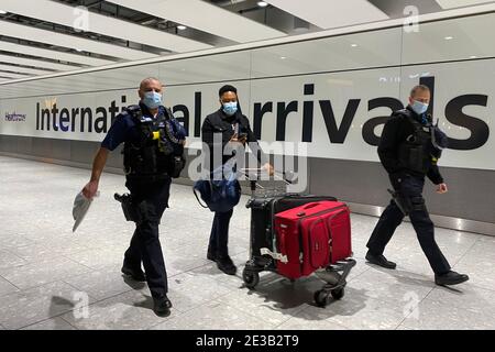 Un passeggero cammina con gli agenti di polizia mentre spinge un tram attraverso la sala arrivi del Terminal 5 all'aeroporto di Heathrow di Londra dopo essere arrivato nel Regno Unito dopo la sospensione dei corridoi di viaggio. I passeggeri che arrivano da qualsiasi luogo al di fuori del Regno Unito, dell'Irlanda, delle Isole della Manica o dell'isola di Man devono avere la prova di un test negativo del coronavirus e di un autoisolamento per 10 giorni. Data immagine: Lunedì 18 gennaio 2021. Foto Stock
