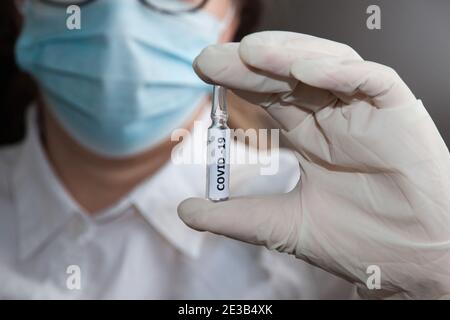 Una donna in una maschera medica e abito da condimento tiene in mano una fiala di vaccino Covid-19. Primo piano Foto Stock