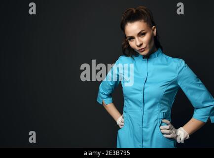 Giovane donna bruna grave infermiera medico in abito medico blu e i guanti in lattice sembrano scettici nei confronti della fotocamera Foto Stock
