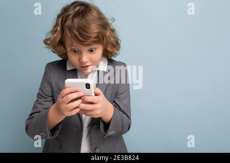 Foto di bel ragazzo scioccato con capelli ricci di colore grigio si adatta tenendo e utilizzando il telefono isolato su sfondo blu guardando sullo smartphone Foto Stock