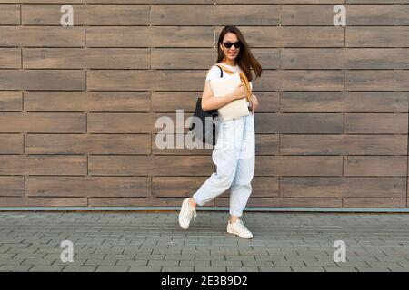 Foto a lunghezza intera di giovane bella brunet beautifyl andando in strada vicino a muro marrone che guarda verso il basso t-shirt bianca e jeans blu chiaro Foto Stock
