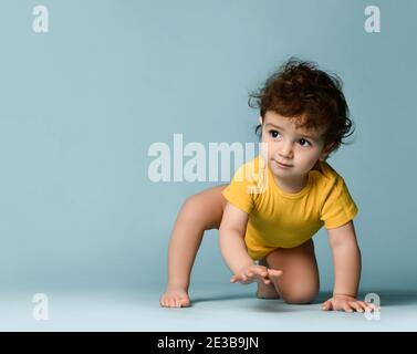 Piccolo bambino carino positivo ricciolo in giallo comodo jumpsuit strisciando sul pavimento e cercando di alzarsi in piedi Foto Stock