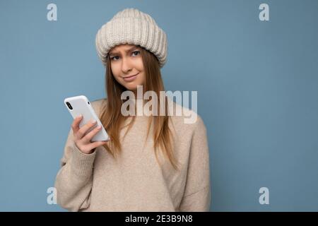 Foto di bella giovane donna bionda sconvolto con elegante beige caldo maglione e maglia inverno caldo cappello beige isolato sopra sfondo blu con Foto Stock