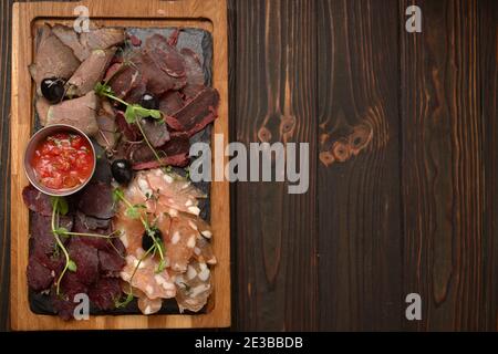 Salumi, salsicce, basturma, affumicati, con salsa e olive, su tavola di legno, su fondo ligneo Foto Stock