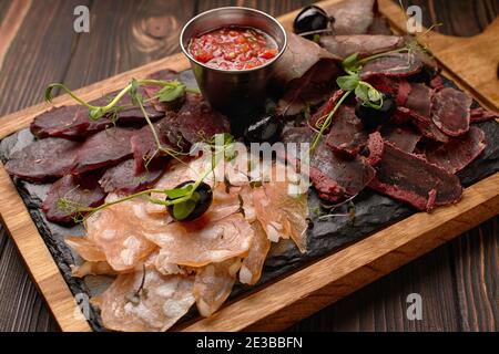 Salumi, salsicce, basturma, affumicati, con salsa e olive, su tavola di legno, su fondo ligneo Foto Stock