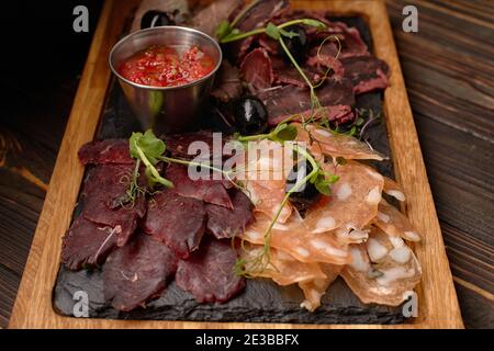 Salumi, salsicce, basturma, affumicati, con salsa e olive, su tavola di legno, su fondo ligneo Foto Stock