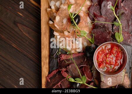 Salumi, salsicce, basturma, affumicati, con salsa e olive, su tavola di legno, su fondo ligneo Foto Stock