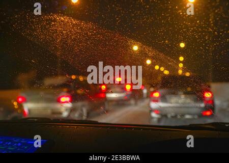 Auto nel traffico in inverno al buio della sera o di notte. Vista attraverso il parabrezza, coperto da gocce d'acqua. Vista sfocata Foto Stock