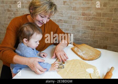 Nonna amorevole, bella donna anziana, che cuoce gustosi biscotti dolci insieme a sua nipote per il giorno di San Valentino, carino piccolo toddler Foto Stock