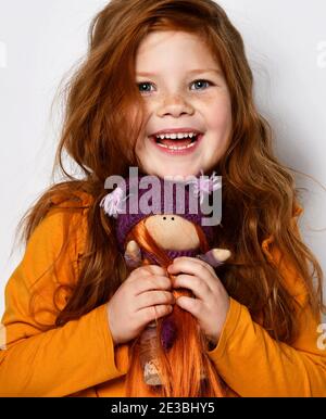 La bambina di cinque anni dai capelli rossi sorride felicemente e tiene una redhaired bambola in mano in un pullover arancione su bianco Foto Stock