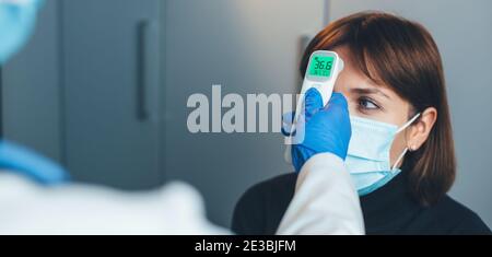 Il medico caucasico con maschera medica sta misurando la temperatura su la fronte del paziente utilizzando un termometro elettronico Foto Stock