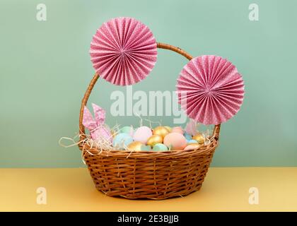 Cesto di vimini con colorate uova di Pasqua in colori pastello e decorato con fiori di carta. Buon biglietto d'auguri pasquale Foto Stock
