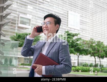 Uomo d'affari indossa giacca, sorridente e parlando al telefono.mentre tenendo un diario. Ambiente urbano. Foto Stock