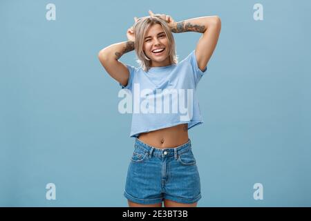 Studio di bella bella bella e gioiosa donna bionda alla moda con tatuaggi sulle armi che si inarricano e sorridono che tengono ampiamente l'indice dita sulla testa fare Foto Stock