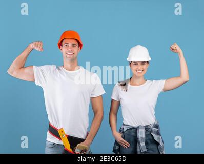 Uomo allegro e donna in caschi protettivi che alzano i pugni, mostrando i muscoli bicipiti Foto Stock