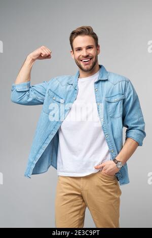 Ritratto di un bell'uomo in una camicia di jeans che lo mostra bicipite con braccio piegato su sfondo grigio Foto Stock