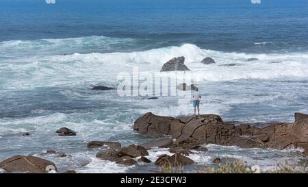 Pescatore in piedi su rocce con canna da pesca, sfondo mare ondulato blu, orizzontale Foto Stock