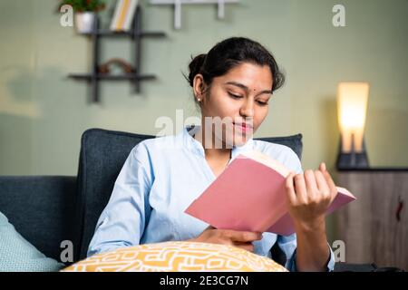 concentrato giovane ragazza leggere libro o studiare durante il tempo libero o per esame a casa da seduta su divano - concetto di hobby di lettura Foto Stock