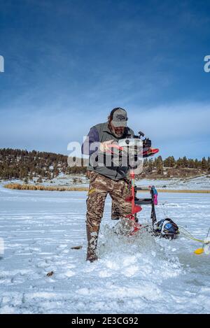 Un uomo in pantaloni camoflauge pratica un buco nel ghiaccio innevato per la pesca sul ghiaccio sul lago artificiale Echo Canyon vicino a Pagosa Springs, Colorado. Foto Stock