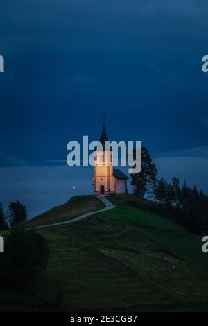Jamnik, Slovenia - ora blu a Jamnik con la chiesa illuminata di San Primoz in cima a una nebbia all'alba. Alpi Giulie sullo sfondo Foto Stock
