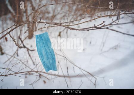 Maschera medica usata appesa su rami in un parco cittadino innevato. Gli spazi pubblici sono sommersi da dispositivi di protezione scartati. Foto Stock