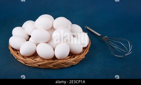 Uova di pollo in cesto di vimini e frusta su sfondo blu Foto Stock
