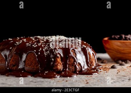 Vista laterale di una torta di cioccolato rotonda con glassa fusa e gocce di cioccolato in una ciotola sullo sfondo. La decorazione di questa deliziosa torta Foto Stock