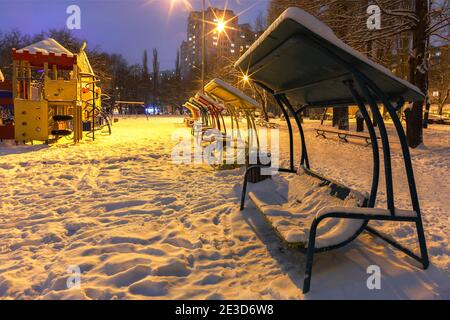 Panchine di legno e un parco giochi nel parco serale della città d'inverno sono coperti di neve sullo sfondo del crepuscolo blu. Foto Stock
