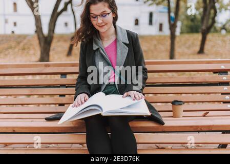 Bella signora indossare occhiali e guardando libro mentre si siede sulla panchina nel parco Foto Stock
