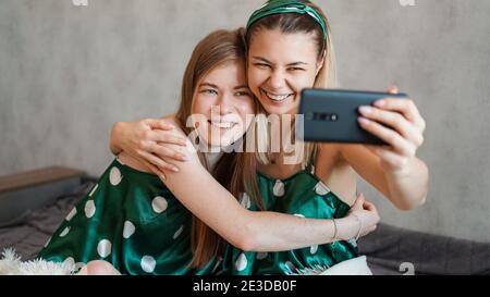 Due bellissime amiche felici che abbracciano e scattano selfie con lo smartphone alla festa di pajama Foto Stock