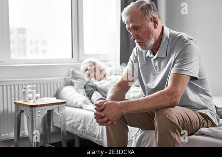 l'uomo pensionato sconvolto si siede nella depressione mentre la sua donna malata che soffre di covid-19, vuole che la moglie sia sana Foto Stock