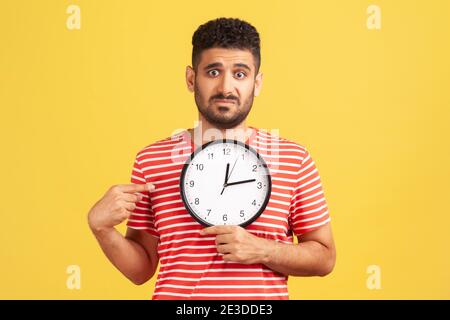 Guardate l'ora! Infelice impaziente ragazzo sopportato in t-shirt a righe che punta grande orologio e l'aspetto dispiaciuto irritato, mostrando orologi a parete a Sbrigati. Foto Stock