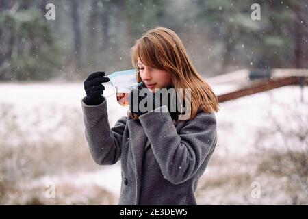 La ragazza caucasica ha rimosso la maschera medica dal viso per bere tè caldo e mantenere caldo all'aperto nella foresta fredda invernale. Foto Stock