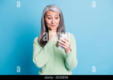 Foto di sorpresa donna anziana guardare in cellulare toccare il mento vestito pullover verde isolato su sfondo blu Foto Stock