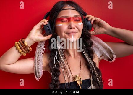 donna indiana con le cuffie, la donna sciamanica ama la musica, ascolta la musica con gli occhi chiusi isolati su sfondo rosso Foto Stock