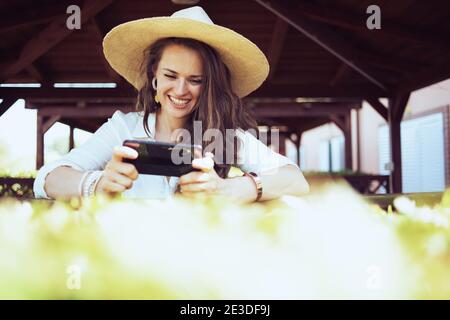 felicemente elegante casalinga in camicia bianca con cappello usando le applicazioni dello smartphone sul ranch. Foto Stock