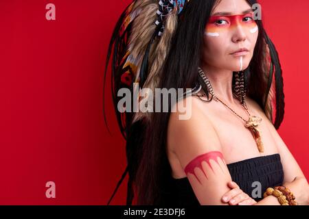fantastico stand modello indiano con braccia incrociate isolate su sfondo rosso, giovane femmina in cima con dipinti sul viso, indossando piume sulla testa, loo Foto Stock