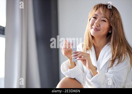 donna si siede sul letto e fa una manicure, usando lima del chiodo, sorride alla macchina fotografica, in accappatoio, a casa Foto Stock