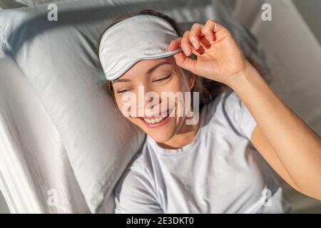 Donna asiatica felice svegliarsi sentendosi rinfrescato dalla maschera di sonno di bellezza occhio che dorme per una buona notte di riposo. Smiing giovane ragazza biraciale a letto. Sano Foto Stock