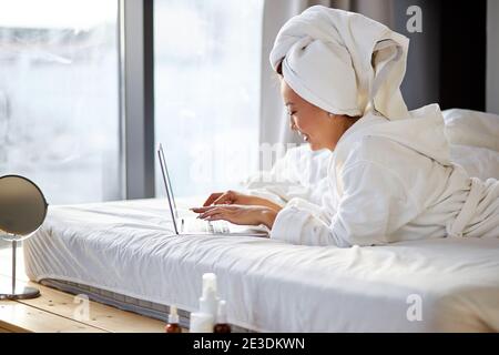 donna asiatica sdraiata sul letto a casa e che lavora sul suo computer portatile, indossando asciugamano e accappatoio, al mattino. lavorando da casa, quarantena coronav Foto Stock