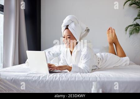 donna asiatica sdraiata sul letto a casa e che lavora sul suo computer portatile, indossando asciugamano e accappatoio, al mattino. lavorando da casa, quarantena coronav Foto Stock