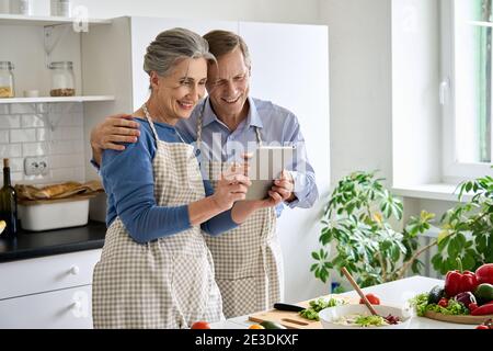 Felice coppia di mezza età preparare l'insalata utilizzando la tavoletta in cucina. Foto Stock