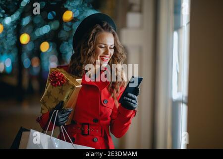 Winter Fun. Donna turistica moderna sorridente in cappotto rosso e berretto nero con borse per lo shopping, regali e smartphone di navigazione negozio on-line fuori della città Foto Stock