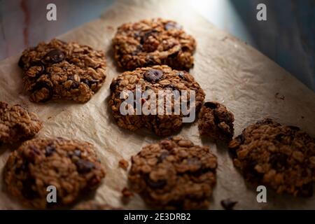 Vista angolata dei biscotti appena fatti sulla carta da forno. Questi sono fatti con avena intera di grano arrotolata con i trucioli di cioccolato scuro. Ci sono pezzi e crumble Foto Stock