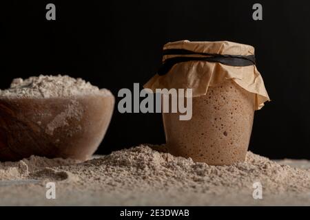 Ingredienti fatti in casa per preparare il pane con una ciotola di farina e una coltura di partenza di pasta di origine in una tazza di vetro coperta con una carta marrone sulla parte superiore Foto Stock