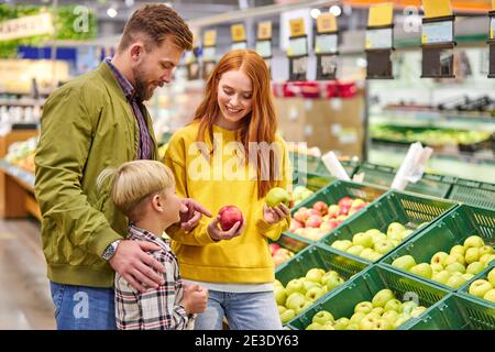 marito e moglie con un capretto comprano frutta, mele. famiglia di tre scegliendo mela fresca nel reparto di frutta del supermercato o del mercato Foto Stock