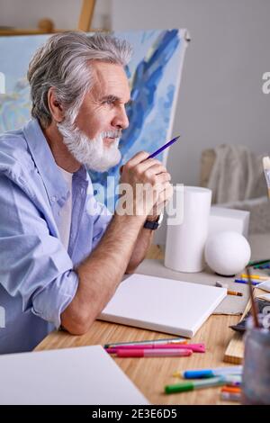 artista dai capelli grigi con matita siedono in contemplazione, nei pensieri. a casa. arte, artigianato, fantasia concetto Foto Stock